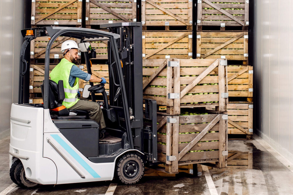 Modern warehouse work on forklift industry and organic agribusiness. Smiling millennial man in helmet in forklift lifts up green apples in container on shelves with many wooden crates | ANG