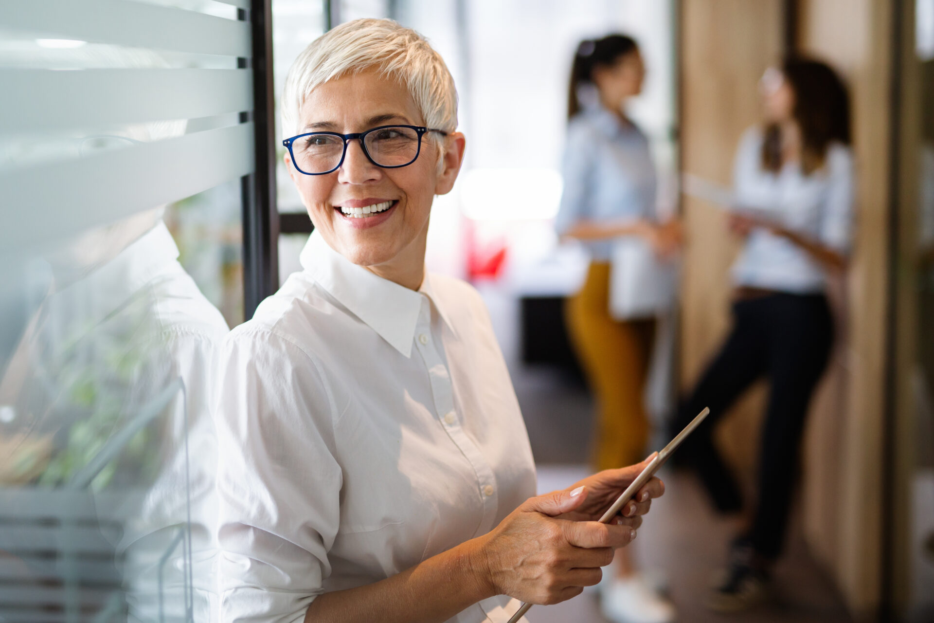 A confident, stylish, mature European businesswoman standing at her desk with a tablet