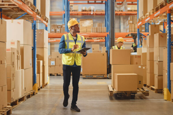 Handsome Male Worker Wearing Hard Hat Holding Digital Tablet Com Handsome Male Worker Wearing Hard Hat Holding Digital Tablet Computer Walking Through Retail Warehouse full of Shelves with Goods. Working in Logistics and Distribution Center.