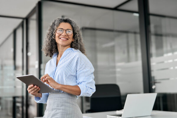 Smiling mature professional business woman bank manager, older happy female executive or lady entrepreneur holding digital tablet pad standing in office at work, looking away at copy space.
