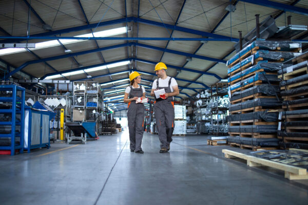 Factory-workers-in-work-wear-and-yellow-helmets-walking-through-industrial-production-hall-and-discussing. Factory-workers-in-work-wear-and-yellow-helmets-walking-through-industrial-production-hall-and-discussing.