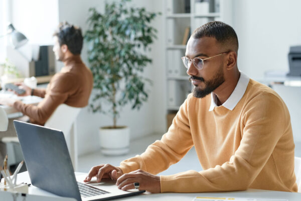 Serious-young-mixed-race-businessman-in-yellow-sweater-working-with-website-on-laptop-in-modern-office